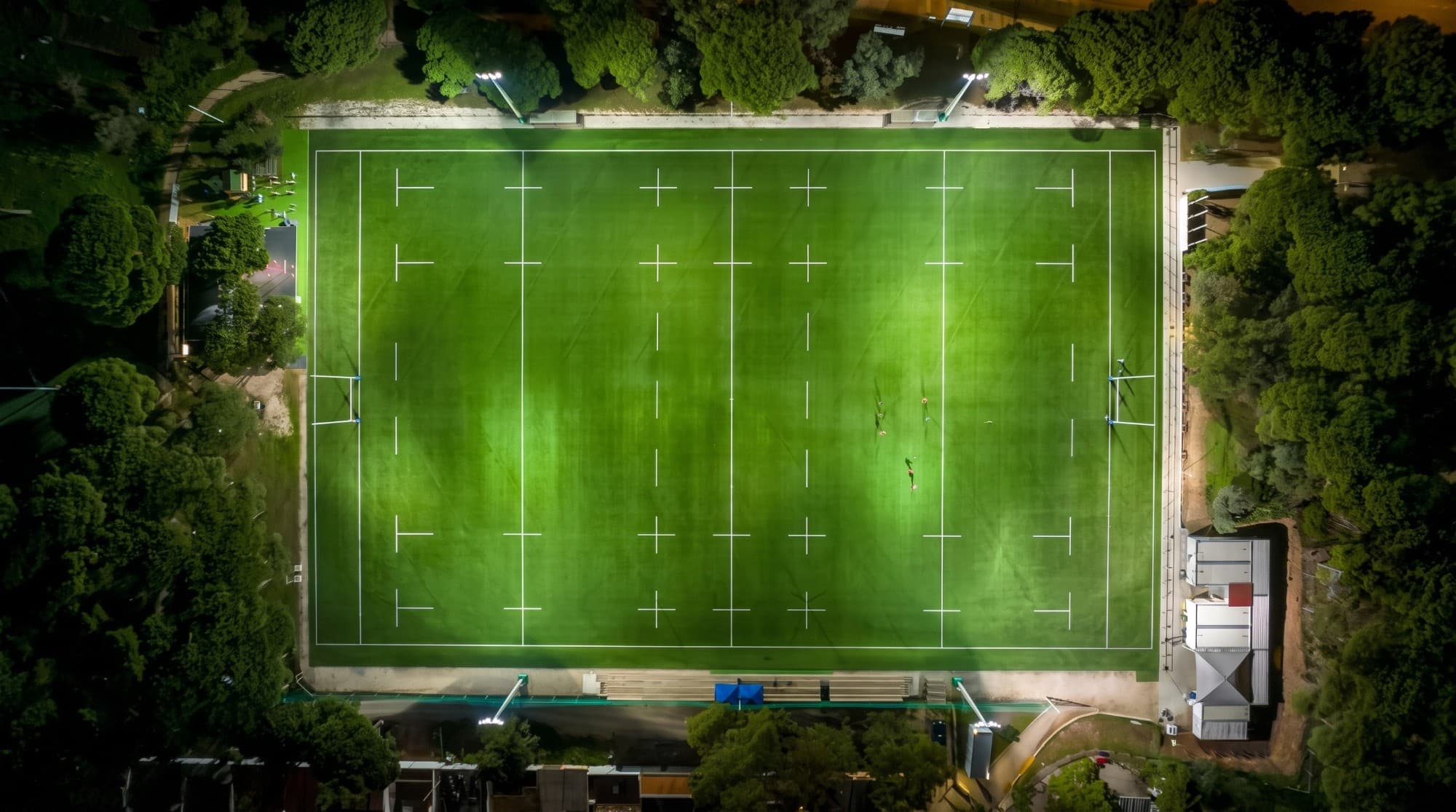 A floodlit school rugby field at night, seen from directly above, surrounded by trees
